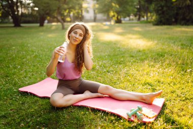 Atletik bir kadın güneşli bir parkta minder üzerinde yoga yapıyor. Spor kıyafetleri içinde güzel bir kadın egzersiz yapıyor, güneş ışınlarında egzersiz yapıyor. Konseptler: fitness, doğa ve vücut bakımı.