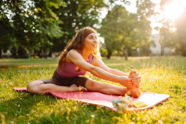 Atletik bir kadın güneşli bir parkta minder üzerinde yoga yapıyor. Spor kıyafetleri içinde güzel bir kadın egzersiz yapıyor, güneş ışınlarında egzersiz yapıyor. Konseptler: fitness, doğa ve vücut bakımı.