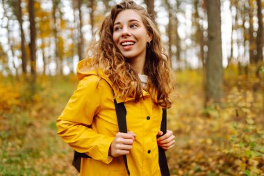 Sarı yağmurluklu genç bir kadın güneşli bir günde sonbahar ormanında gezinir. Neşeli bir sırt çantası güz mevsiminin güzelliğinin tadını çıkarır. Macera ve mevsimlik kavramı.