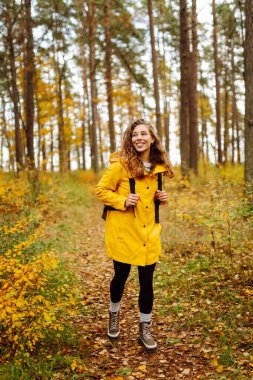 Sarı yağmurluklu genç bir kadın güneşli bir günde sonbahar ormanında gezinir. Neşeli bir sırt çantası güz mevsiminin güzelliğinin tadını çıkarır. Macera ve mevsimlik kavramı.