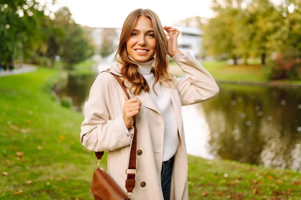 Şık bir trençkot giymiş neşeli bir kadın sonbahar parkında poz veriyor. Güzel bir kadın dışarıda yürümekten hoşlanır. Güzellik ve moda konsepti. Sonbahar havası.