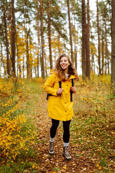 Sarı yağmurluklu genç bir kadın güneşli bir günde sonbahar ormanında gezinir. Neşeli bir sırt çantası güz mevsiminin güzelliğinin tadını çıkarır. Macera ve mevsimlik kavramı.