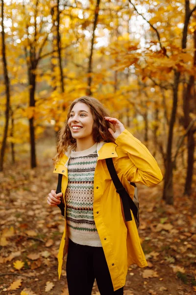 Sarı yağmurluklu genç bir kadın güneşli bir günde sonbahar ormanında gezinir. Neşeli bir sırt çantası güz mevsiminin güzelliğinin tadını çıkarır. Macera ve mevsimlik kavramı.