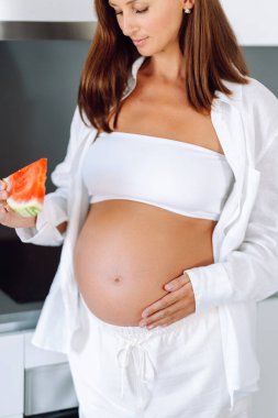 Müstakbel bir anne modern bir mutfakta elinde bir dilim karpuz tutuyor. Hamile bir kadın sıcak bir odada karpuz yiyor. Sağlıklı beslenme. Annelik ve bakım kavramları.