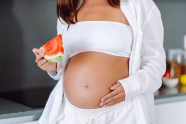 Müstakbel bir anne modern bir mutfakta elinde bir dilim karpuz tutuyor. Hamile bir kadın sıcak bir odada karpuz yiyor. Sağlıklı beslenme. Annelik ve bakım kavramları.