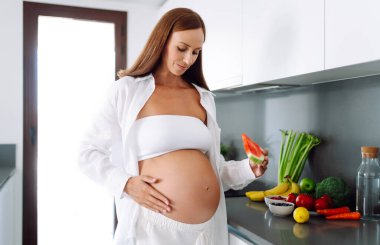 Müstakbel bir anne modern bir mutfakta elinde bir dilim karpuz tutuyor. Hamile bir kadın sıcak bir odada karpuz yiyor. Sağlıklı beslenme. Annelik ve bakım kavramları.