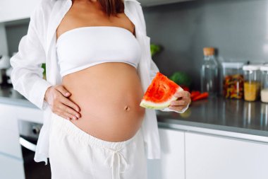 Müstakbel bir anne modern bir mutfakta elinde bir dilim karpuz tutuyor. Hamile bir kadın sıcak bir odada karpuz yiyor. Sağlıklı beslenme. Annelik ve bakım kavramları.