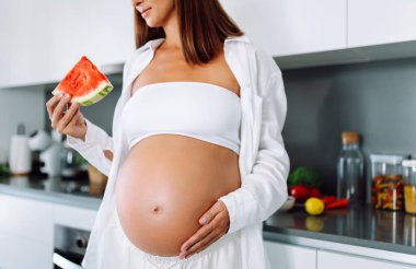Müstakbel bir anne modern bir mutfakta elinde bir dilim karpuz tutuyor. Hamile bir kadın sıcak bir odada karpuz yiyor. Sağlıklı beslenme. Annelik ve bakım kavramları.
