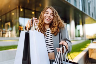 Alışveriş çantalı neşeli bir kadın modern bir alışveriş merkezinin yakınında eğleniyor. Güzel bir kadın, alışverişlerinin çantalarla yapılmasının keyfini çıkarır. Tüketim kavramı, indirimler.