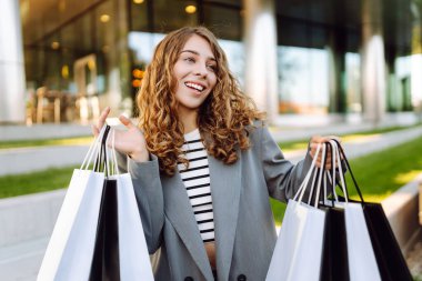 Alışveriş çantalı neşeli bir kadın modern bir alışveriş merkezinin yakınında eğleniyor. Güzel bir kadın, alışverişlerinin çantalarla yapılmasının keyfini çıkarır. Tüketim kavramı, indirimler.