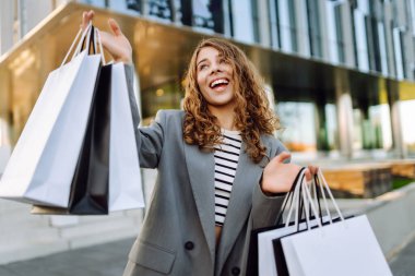 Alışveriş çantalı neşeli bir kadın modern bir alışveriş merkezinin yakınında eğleniyor. Güzel bir kadın, alışverişlerinin çantalarla yapılmasının keyfini çıkarır. Tüketim kavramı, indirimler.