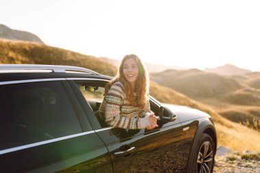 Mutlu bir gezgin gün batımında arabanın camından sarkar. Genç kadın kendini özgür hissediyor ve araba sürerken dağ manzarasının tadını çıkarıyor. Macera ve ulaşım konsepti. Etkin yaşam biçimi.