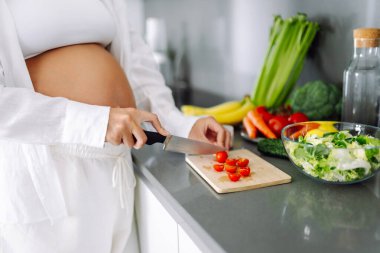 Genç hamile bir kadın modern bir mutfakta dururken kereviz doğruyor. Müstakbel güzel bir anne sıcak bir odada taze meyve ve sebze seçer. Sağlıklı yaşam tarzı, beslenme, yiyecek kavramları.