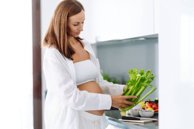 Genç hamile bir kadın modern bir mutfakta dururken kereviz doğruyor. Müstakbel güzel bir anne sıcak bir odada taze meyve ve sebze seçer. Sağlıklı yaşam tarzı, beslenme, yiyecek kavramları.