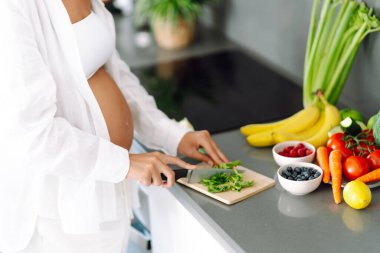 Genç hamile bir kadın modern bir mutfakta dururken kereviz doğruyor. Müstakbel güzel bir anne sıcak bir odada taze meyve ve sebze seçer. Sağlıklı yaşam tarzı, beslenme, yiyecek kavramları.