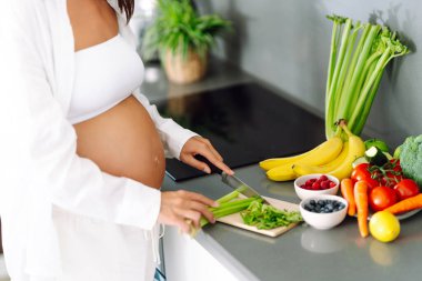 Genç hamile bir kadın modern bir mutfakta dururken kereviz doğruyor. Müstakbel güzel bir anne sıcak bir odada taze meyve ve sebze seçer. Sağlıklı yaşam tarzı, beslenme, yiyecek kavramları.