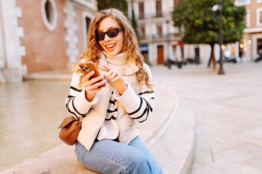 Güneş gözlüklü genç bir kadın telefonuyla şehrin güneşli sokaklarında geziniyor. Bir turist gün batımının tadını çıkarıyor, telefonundan sosyal medyayı kontrol ediyor. Konsept: Turizm, yürüyüş.