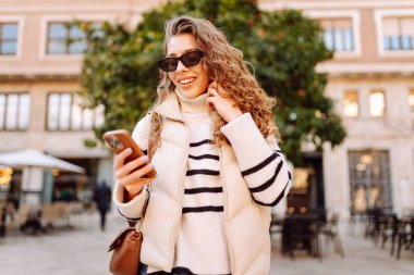 Güneş gözlüklü genç bir kadın telefonuyla şehrin güneşli sokaklarında geziniyor. Bir turist gün batımının tadını çıkarıyor, telefonundan sosyal medyayı kontrol ediyor. Konsept: Turizm, yürüyüş.