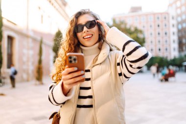 Güneş gözlüklü genç bir kadın telefonuyla şehrin güneşli sokaklarında geziniyor. Bir turist gün batımının tadını çıkarıyor, telefonundan sosyal medyayı kontrol ediyor. Konsept: Turizm, yürüyüş.