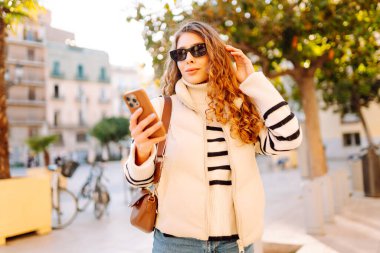 Güneş gözlüklü genç bir kadın telefonuyla şehrin güneşli sokaklarında geziniyor. Bir turist gün batımının tadını çıkarıyor, telefonundan sosyal medyayı kontrol ediyor. Konsept: Turizm, yürüyüş.