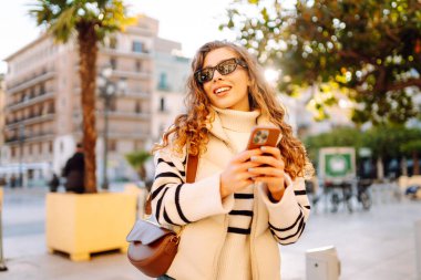 Güneş gözlüklü genç bir kadın telefonuyla şehrin güneşli sokaklarında geziniyor. Bir turist gün batımının tadını çıkarıyor, telefonundan sosyal medyayı kontrol ediyor. Konsept: Turizm, yürüyüş.