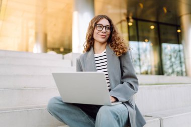 Şık giyinmiş bir kadın basamaklarda dizüstü bilgisayarla modern bir binanın arka planına karşı oturur. Güzel bayan serbest yazar dizüstü bilgisayarda çalışıyor ve güneşli günün tadını çıkarıyor. Uzak iş ve iş için kavram.