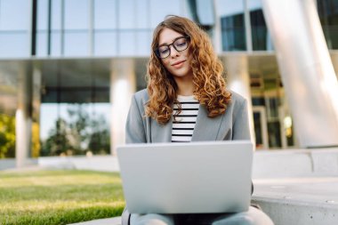 Şık giyinmiş bir kadın basamaklarda dizüstü bilgisayarla modern bir binanın arka planına karşı oturur. Güzel bayan serbest yazar dizüstü bilgisayarda çalışıyor ve güneşli günün tadını çıkarıyor. Uzak iş ve iş için kavram.