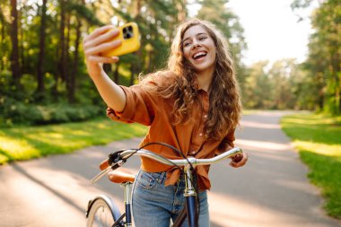 Selfie zamanı! Telefonlu neşeli kadın güneşli bir parkta bisikletle otururken selfie çekiyor. Genç kadın güneşli bir günün tadını çıkarıyor ve bisiklete biniyor. Rahatlama, blog yazma ve hafta sonları için kavramlar.