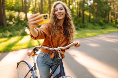 Selfie zamanı! Telefonlu neşeli kadın güneşli bir parkta bisikletle otururken selfie çekiyor. Genç kadın güneşli bir günün tadını çıkarıyor ve bisiklete biniyor. Rahatlama, blog yazma ve hafta sonları için kavramlar.