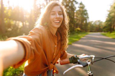 Selfie zamanı! Telefonlu neşeli kadın güneşli bir parkta bisikletle otururken selfie çekiyor. Genç kadın güneşli bir günün tadını çıkarıyor ve bisiklete biniyor. Rahatlama, blog yazma ve hafta sonları için kavramlar.