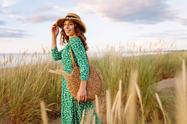 Uzun hasır şapkalı gülümseyen bir kadın sahile yakın poz veriyor. Saman çantalı güzel bir kadın deniz manzarasının ve gün batımının tadını çıkarıyor. Kavram: doğa, güzellik, yaşam tarzı.