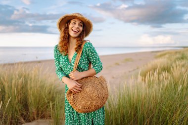 Uzun hasır şapkalı gülümseyen bir kadın sahile yakın poz veriyor. Saman çantalı güzel bir kadın deniz manzarasının ve gün batımının tadını çıkarıyor. Kavram: doğa, güzellik, yaşam tarzı.