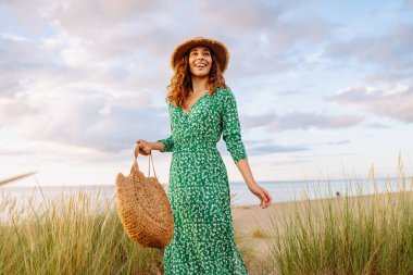 Uzun hasır şapkalı gülümseyen bir kadın sahile yakın poz veriyor. Saman çantalı güzel bir kadın deniz manzarasının ve gün batımının tadını çıkarıyor. Kavram: doğa, güzellik, yaşam tarzı.