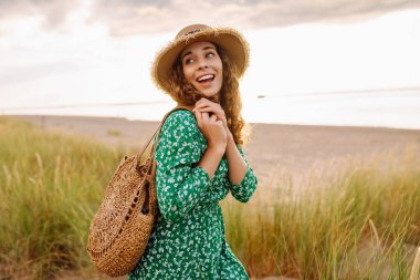 Uzun hasır şapkalı gülümseyen bir kadın sahile yakın poz veriyor. Saman çantalı güzel bir kadın deniz manzarasının ve gün batımının tadını çıkarıyor. Kavram: doğa, güzellik, yaşam tarzı.