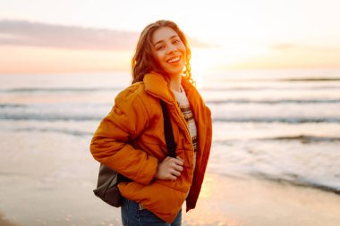 Sırt çantalı güzel bir kadın şafak vakti kıyıda geziniyor. Gezgin güneş ışınlarındaki deniz manzarasının tadını çıkarıyor. Seyahat, keyif ve özgürlük kavramları.