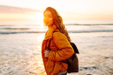 Sırt çantalı güzel bir kadın şafak vakti kıyıda geziniyor. Gezgin güneş ışınlarındaki deniz manzarasının tadını çıkarıyor. Seyahat, keyif ve özgürlük kavramları.