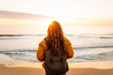 Sırt çantalı güzel bir kadın şafak vakti kıyıda geziniyor. Gezgin güneş ışınlarındaki deniz manzarasının tadını çıkarıyor. Seyahat, keyif ve özgürlük kavramları.