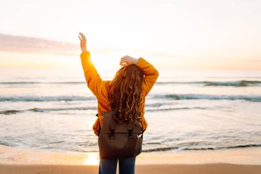 Sırt çantalı güzel bir kadın şafak vakti kıyıda geziniyor. Gezgin güneş ışınlarındaki deniz manzarasının tadını çıkarıyor. Seyahat, keyif ve özgürlük kavramları.