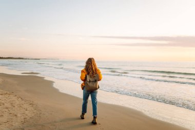 Sırt çantalı güzel bir kadın şafak vakti kıyıda geziniyor. Gezgin güneş ışınlarındaki deniz manzarasının tadını çıkarıyor. Seyahat, keyif ve özgürlük kavramları.