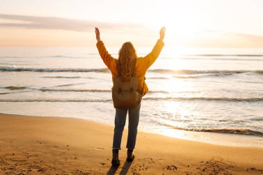 Sırt çantalı güzel bir kadın şafak vakti kıyıda geziniyor. Gezgin güneş ışınlarındaki deniz manzarasının tadını çıkarıyor. Seyahat, keyif ve özgürlük kavramları.