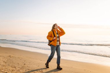 Sırt çantalı güzel bir kadın şafak vakti kıyıda geziniyor. Gezgin güneş ışınlarındaki deniz manzarasının tadını çıkarıyor. Seyahat, keyif ve özgürlük kavramları.