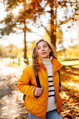 Sırt çantası ve parlak ceketli genç bir gezginin portresi sonbahar parkında gün batımında geziniyor. Mutlu kadın ormanda eğleniyor ve güneşli bir günün tadını çıkarıyor. Seyahat ve hafta sonu kavramı.