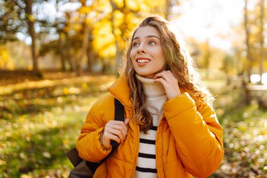 Sırt çantası ve parlak ceketli genç bir gezginin portresi sonbahar parkında gün batımında geziniyor. Mutlu kadın ormanda eğleniyor ve güneşli bir günün tadını çıkarıyor. Seyahat ve hafta sonu kavramı.