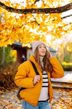 Sırt çantası ve parlak ceketli genç bir gezginin portresi sonbahar parkında gün batımında geziniyor. Mutlu kadın ormanda eğleniyor ve güneşli bir günün tadını çıkarıyor. Seyahat ve hafta sonu kavramı.