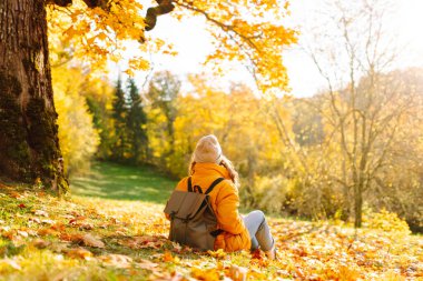 Sırt çantası ve parlak ceketli genç bir gezginin portresi sonbahar parkında gün batımında geziniyor. Mutlu kadın ormanda eğleniyor ve güneşli bir günün tadını çıkarıyor. Seyahat ve hafta sonu kavramı.