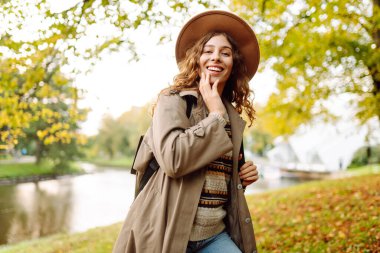Sonbahar parkında yağmurluk ve şapkalı neşeli bir kadın bir nehrin kenarında duruyor. Sırt çantalı genç bir turist doğa ve doğa yürüyüşünün tadını çıkarıyor. Mevsimlik ve turizm kavramı.
