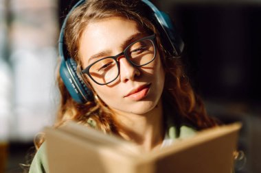 Kablosuz kulaklık takan ve güneş ışığında kitap tutan genç bir kadının yakın çekimi. Gözlüklü, gülümseyen bir kadın kitap okuyor ve rahat bir odada müzik dinlemekten hoşlanıyor. Konsept: eğitim, teknoloji.
