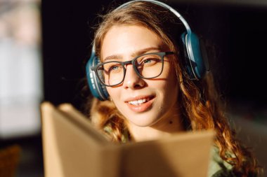 Kablosuz kulaklık takan ve güneş ışığında kitap tutan genç bir kadının yakın çekimi. Gözlüklü, gülümseyen bir kadın kitap okuyor ve rahat bir odada müzik dinlemekten hoşlanıyor. Konsept: eğitim, teknoloji.