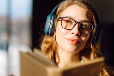 Kablosuz kulaklık takan ve güneş ışığında kitap tutan genç bir kadının yakın çekimi. Gözlüklü, gülümseyen bir kadın kitap okuyor ve rahat bir odada müzik dinlemekten hoşlanıyor. Konsept: eğitim, teknoloji.