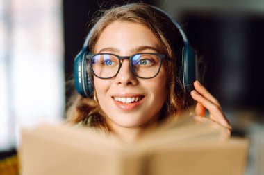 Kablosuz kulaklık takan ve güneş ışığında kitap tutan genç bir kadının yakın çekimi. Gözlüklü, gülümseyen bir kadın kitap okuyor ve rahat bir odada müzik dinlemekten hoşlanıyor. Konsept: eğitim, teknoloji.